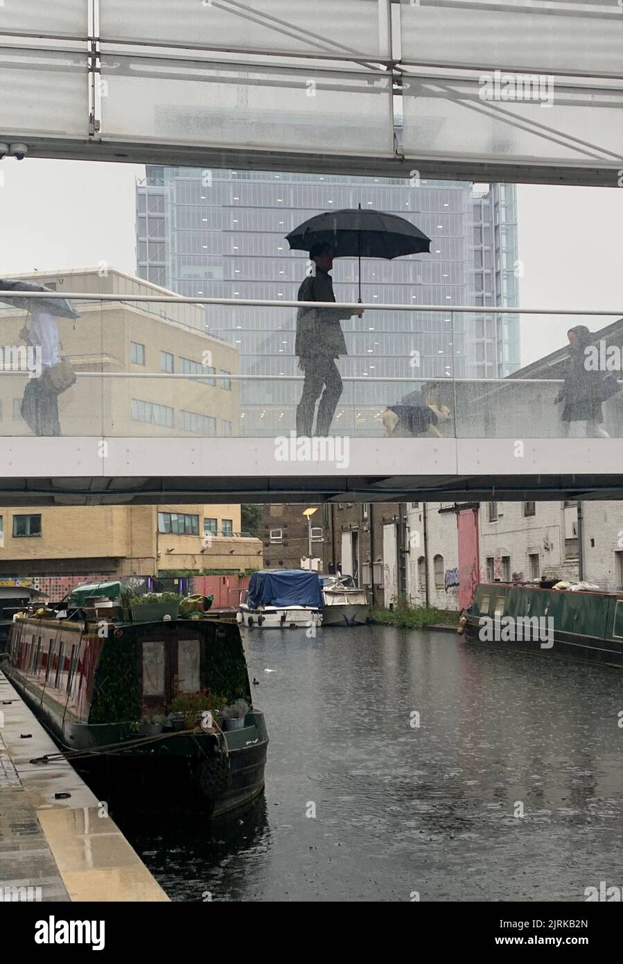 Les voyageurs se réfugient sous des parasols en traversant un pont sur le canal de Paddington, dans l'ouest de Londres, pendant une averse de pluie. Le bureau du met a émis un avertissement jaune pour les orages et les fortes pluies dans le sud et l'est de l'Angleterre, les conditions de conduite pouvant être affectées par les projections d'eau, les eaux stagnantes et même la grêle, ainsi que les retards possibles dans les services ferroviaires, les coupures d'électricité, les inondations et les éclairs. Date de la photo: Jeudi 25 août 2022. Banque D'Images