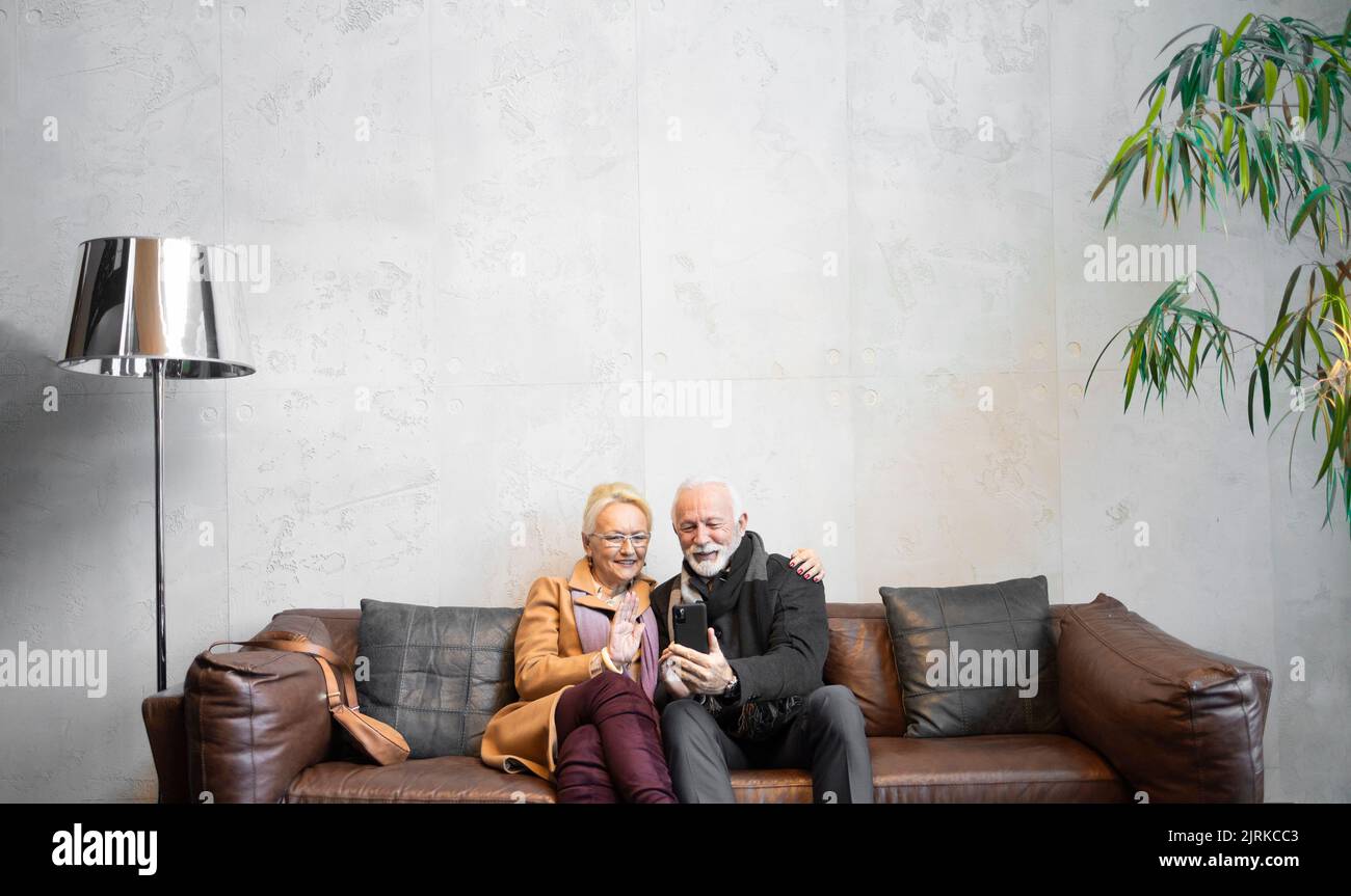 Couple âgé assis dans le hall de l'hôtel et parlant à leurs amis et à leur famille par vidéoconférence Banque D'Images