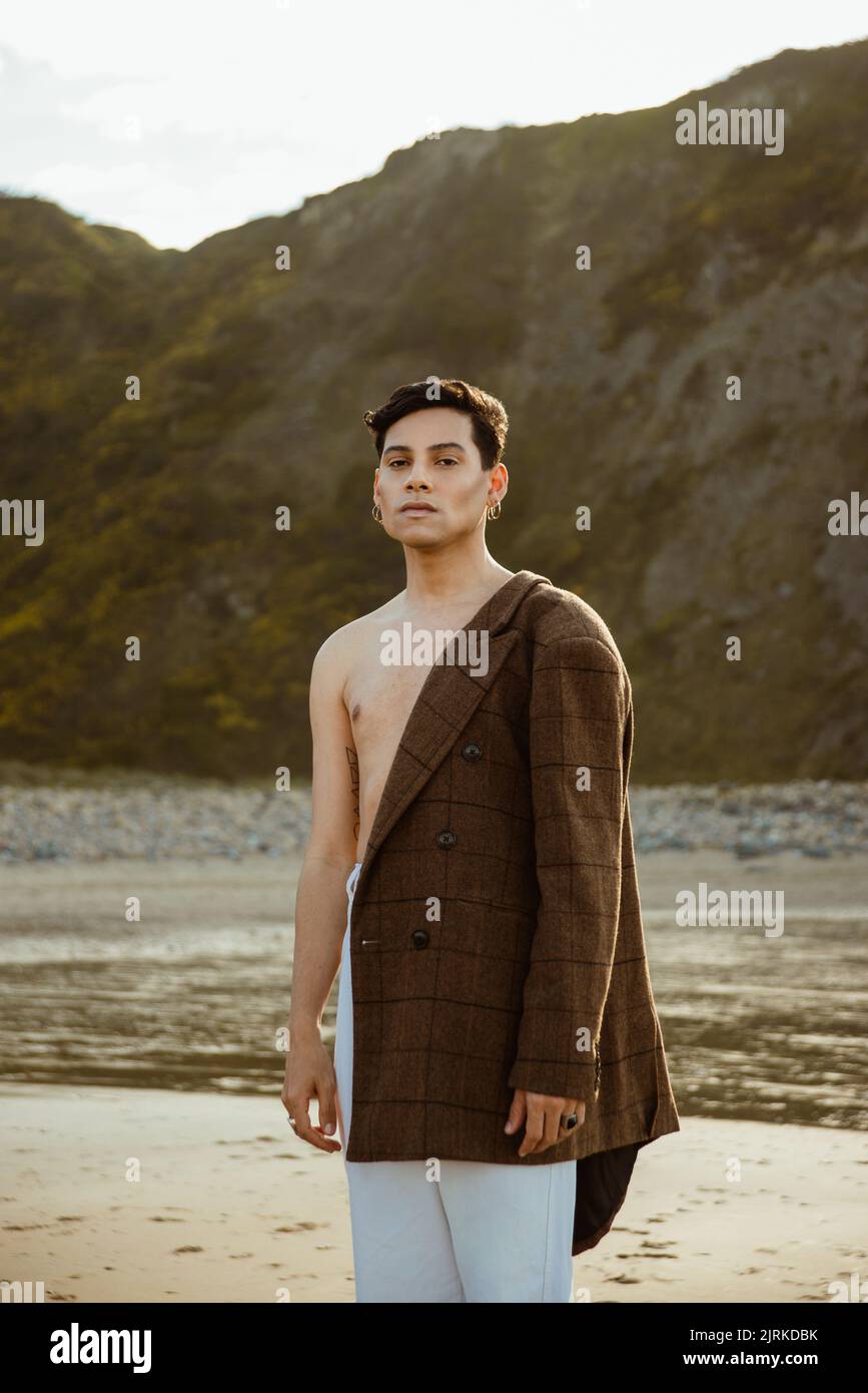Jeune homme ethnique regardant la caméra et mettant une veste élégante tout en se relaxant sur une plage de sable près de la mer et de la falaise en été Banque D'Images