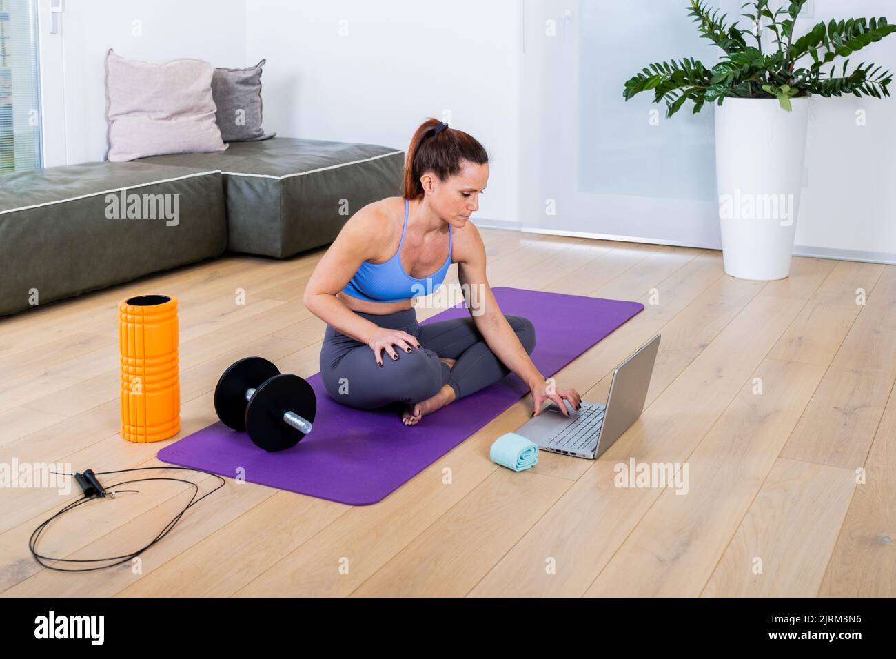 Placez la jeune femme dans des vêtements de sport à la recherche d'un cours de vidéo d'entraînement sur le Web à l'aide d'un ordinateur portable, allongé sur un tapis de yoga dans le salon moderne Banque D'Images
