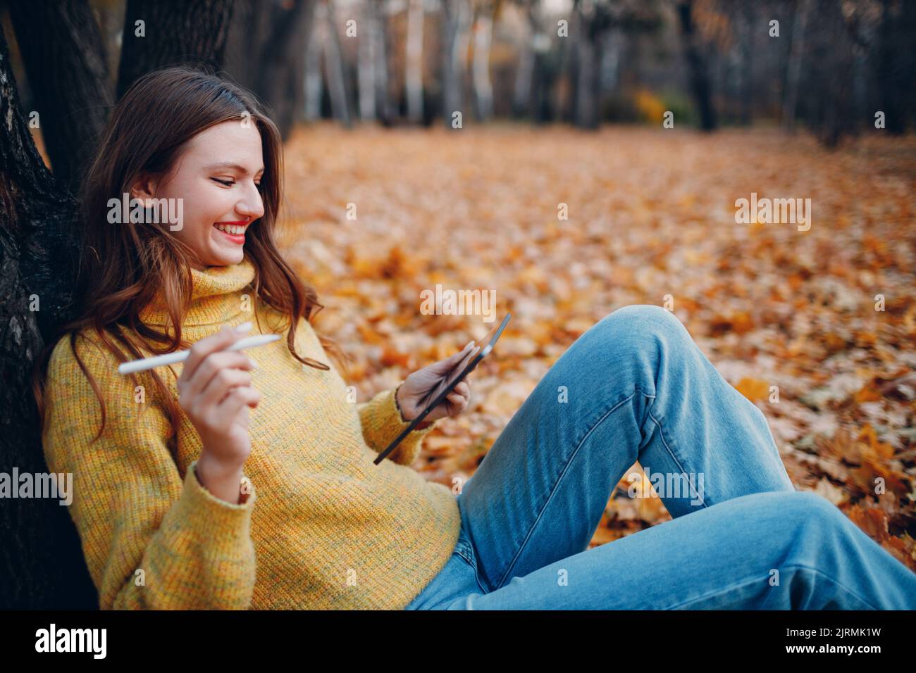 Jeune femme modèle assis avec un PC tablette et peinture avec stylet dans le parc d'automne avec feuilles d'érable à feuillage jaune. Banque D'Images