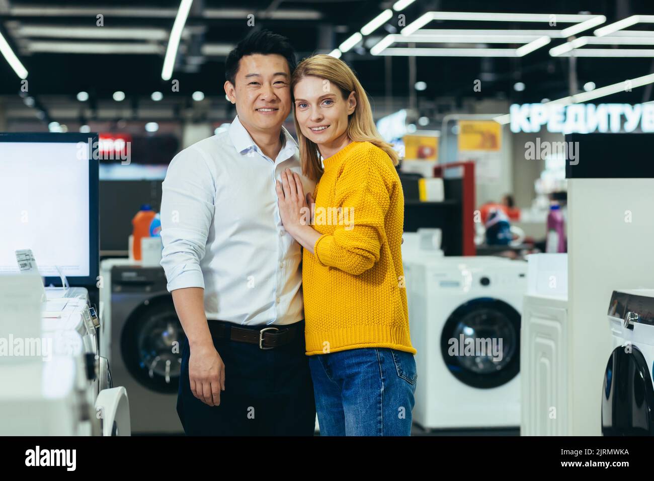 Portrait d'un jeune couple marié magasiner dans un supermarché d'appareils ménagers, une famille diversifiée d'un homme et d'une femme souriant et regardant l'appareil photo, le choix et l'achat d'appareils électroménagers Banque D'Images