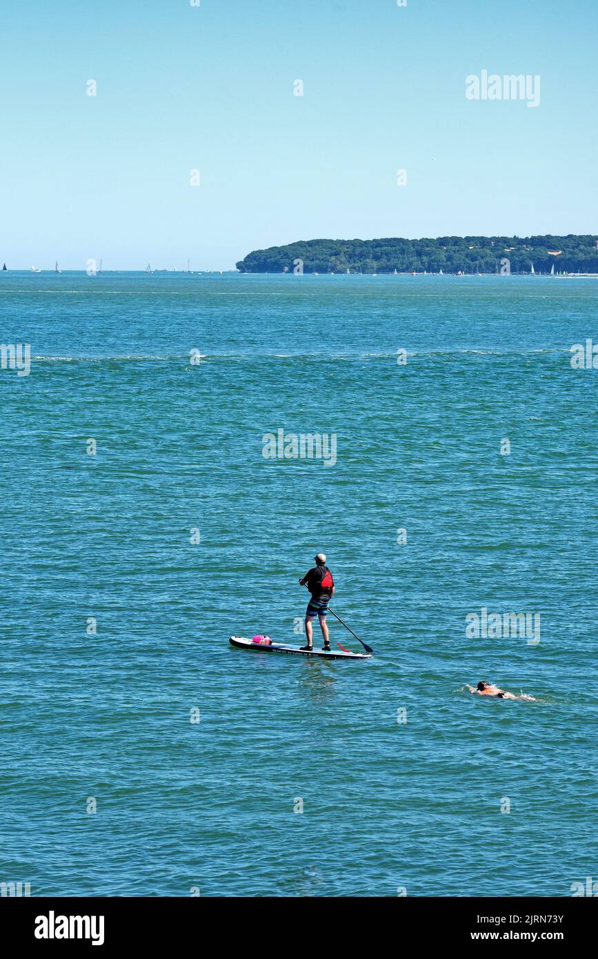 Un homme pagayer avec un nageur à côté de lui sur la mer bleue à Lepe le jour de l'été Hampshire Angleterre Royaume-Uni Banque D'Images
