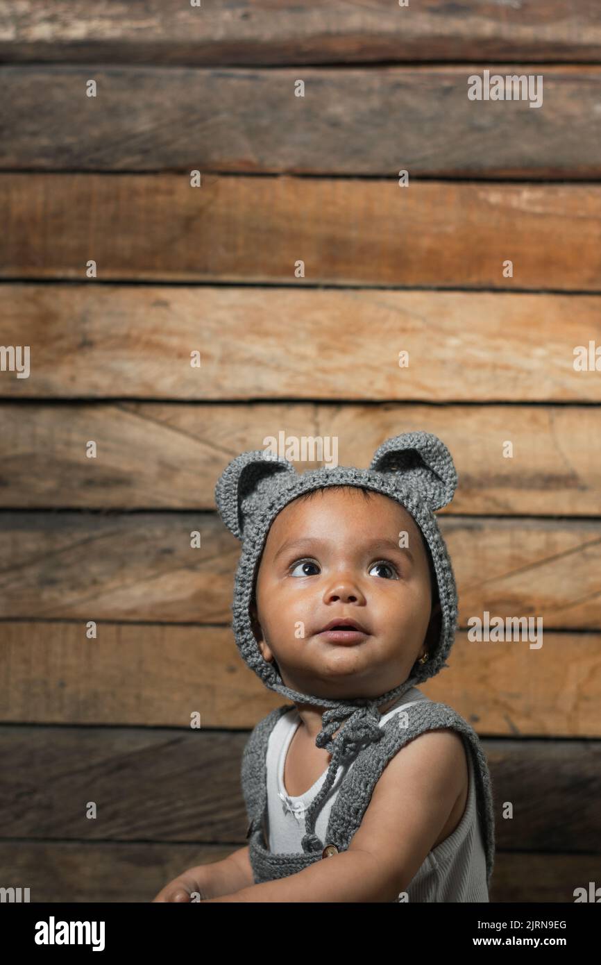 belle latina bébé avec peau brune, portant une combinaison et un chapeau avec des oreilles de souris, sur un fond en bois. regardant curieusement vers le haut, la recherche d'elle Banque D'Images