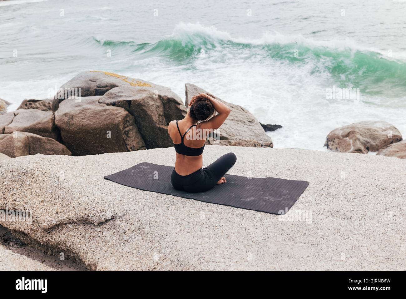 Vue arrière d'une jeune femme assise sur un tapis et réchauffant son cou par l'océan. Femme faisant du yoga et regardant sur les vagues. Banque D'Images