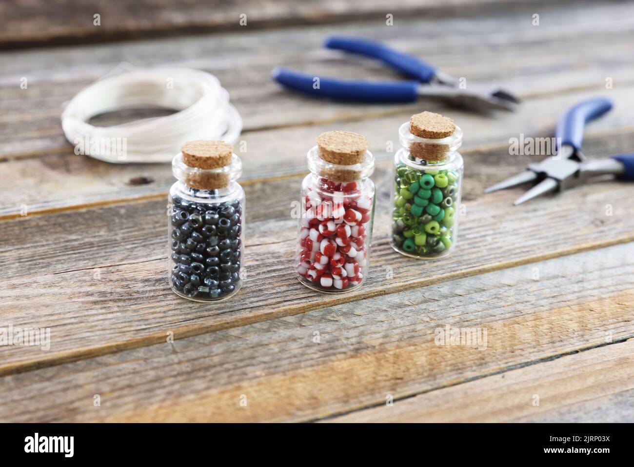 Perles de graines rouges, vertes et noires dans de petits pots sur une surface en bois avec coupe-fils, pinces et cordelette en arrière-plan. Banque D'Images