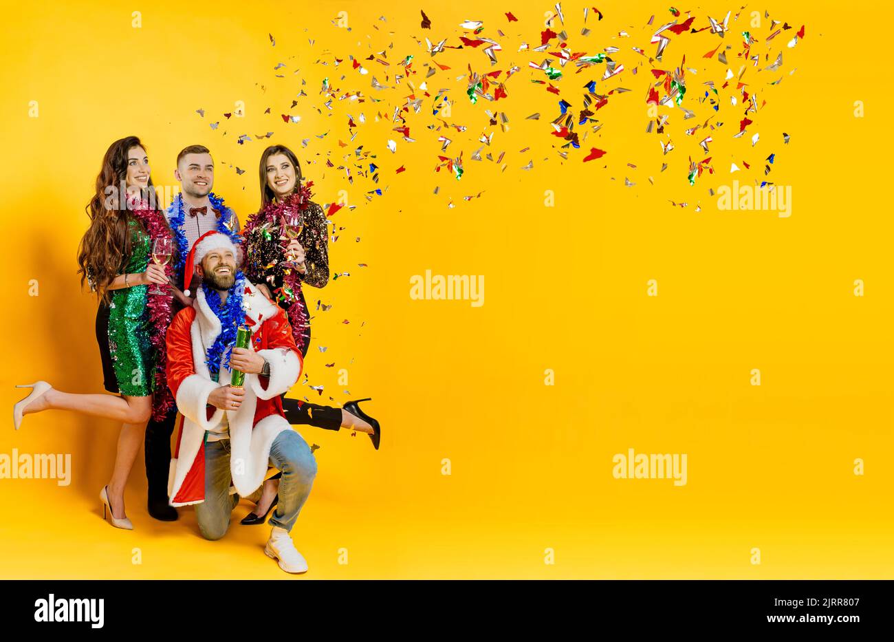 Fête confettis. Un groupe d'amis dans la guirlande du nouvel an et un costume de santa photographiez un coqueliver confetti sur un fond jaune isolé avec espace de copie. B Banque D'Images