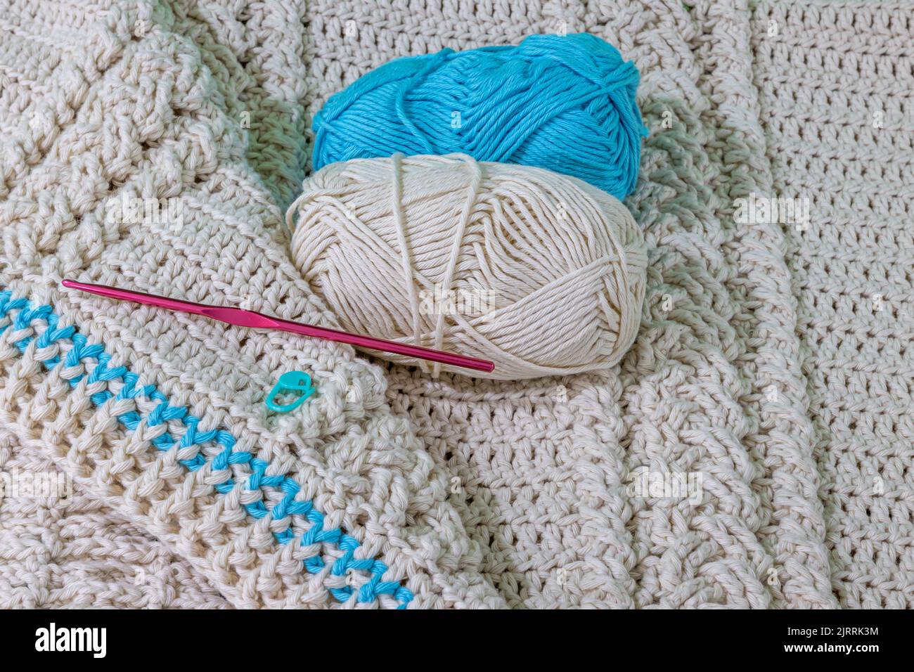 Une pelote beige et une pelote bleue, un crochet en métal rose et un marqueur à point vert sur une couverture en crochet beige en fil de coton, des tresses entrelacées, une cro originale relevée Banque D'Images