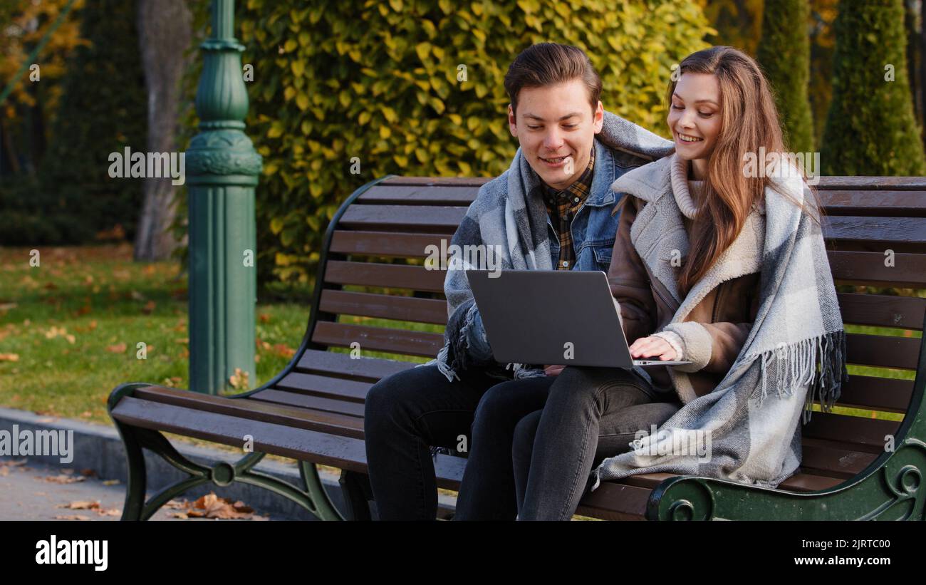 Couple indépendants deux partenaires personnes jeunes femmes et hommes entrepreneurs caucasiens assis sur banc dans le parc utiliser le wifi urbain travail sur ordinateur portable regarder l'appareil Banque D'Images