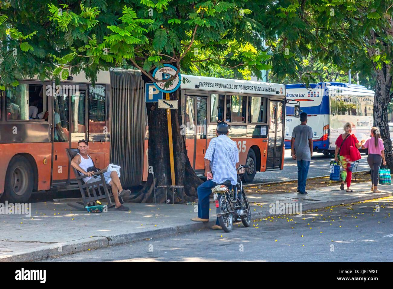 Une personne cubaine est assise dans une chaise berçante à côté d'un arrêt de bus parlant avec un homme dans un cycle. D'autres personnes marchant dans la région. Banque D'Images