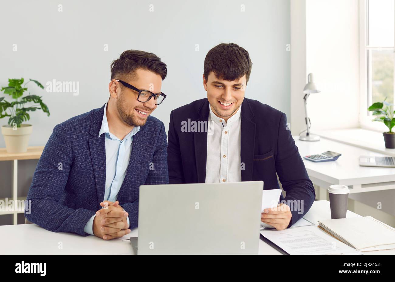 Deux jeunes hommes d'affaires heureux travaillant au bureau et utilisant un ordinateur portable ensemble Banque D'Images
