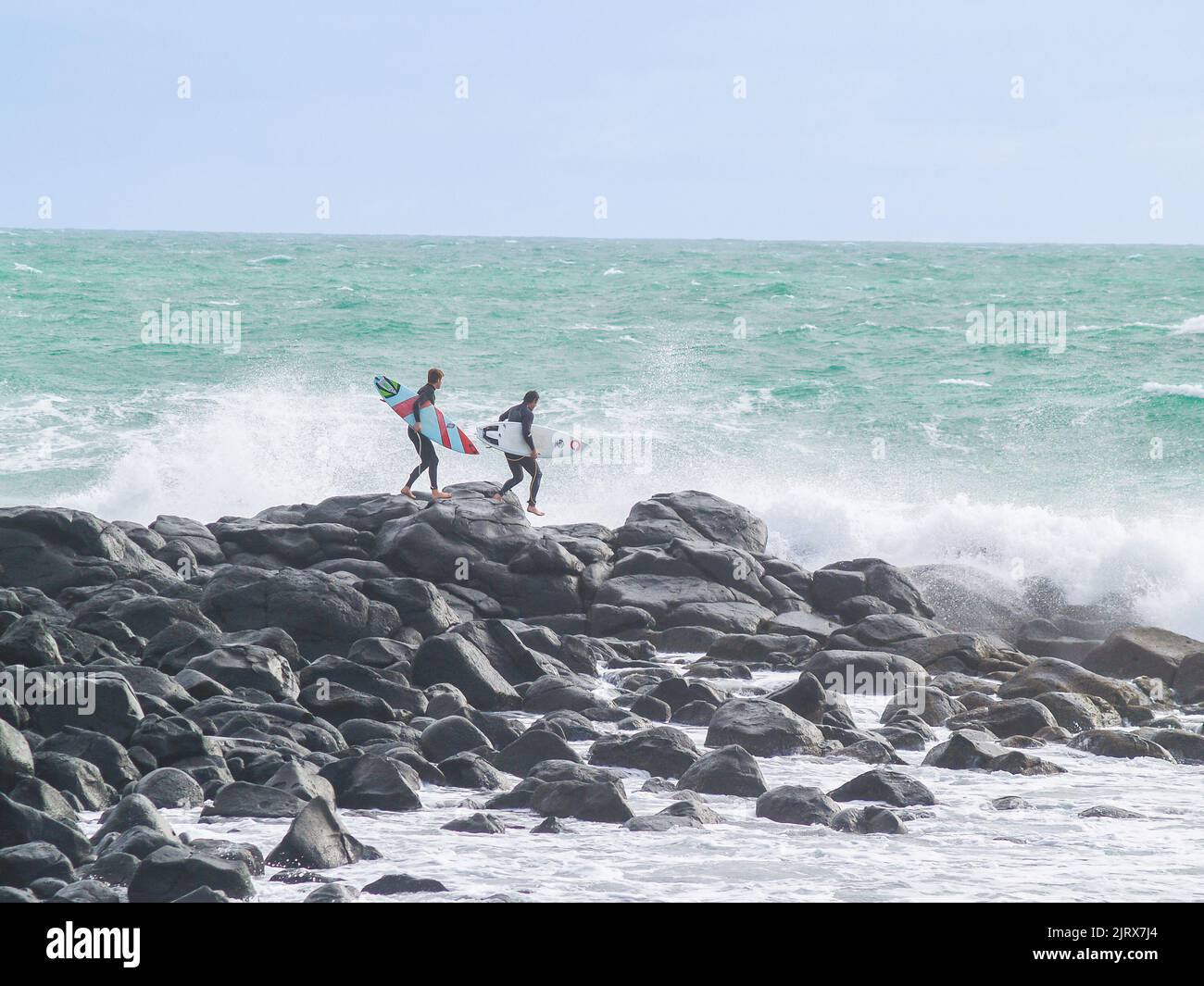 Raglan, Nouvelle-Zélande - 7 juin 2008 de jeunes surfeurs mâles clamberent sur le rivage rocheux pour surfer alors que d'énormes vagues se brisent autour de lui. Banque D'Images
