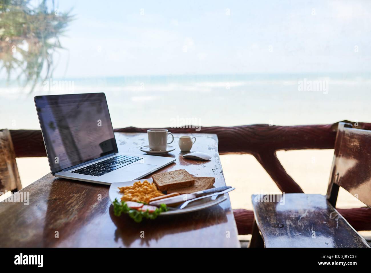Petit déjeuner avec la meilleure vue. Un ordinateur portable et un petit déjeuner fraîchement préparé sur une table avec vue sur la plage en arrière-plan. Banque D'Images