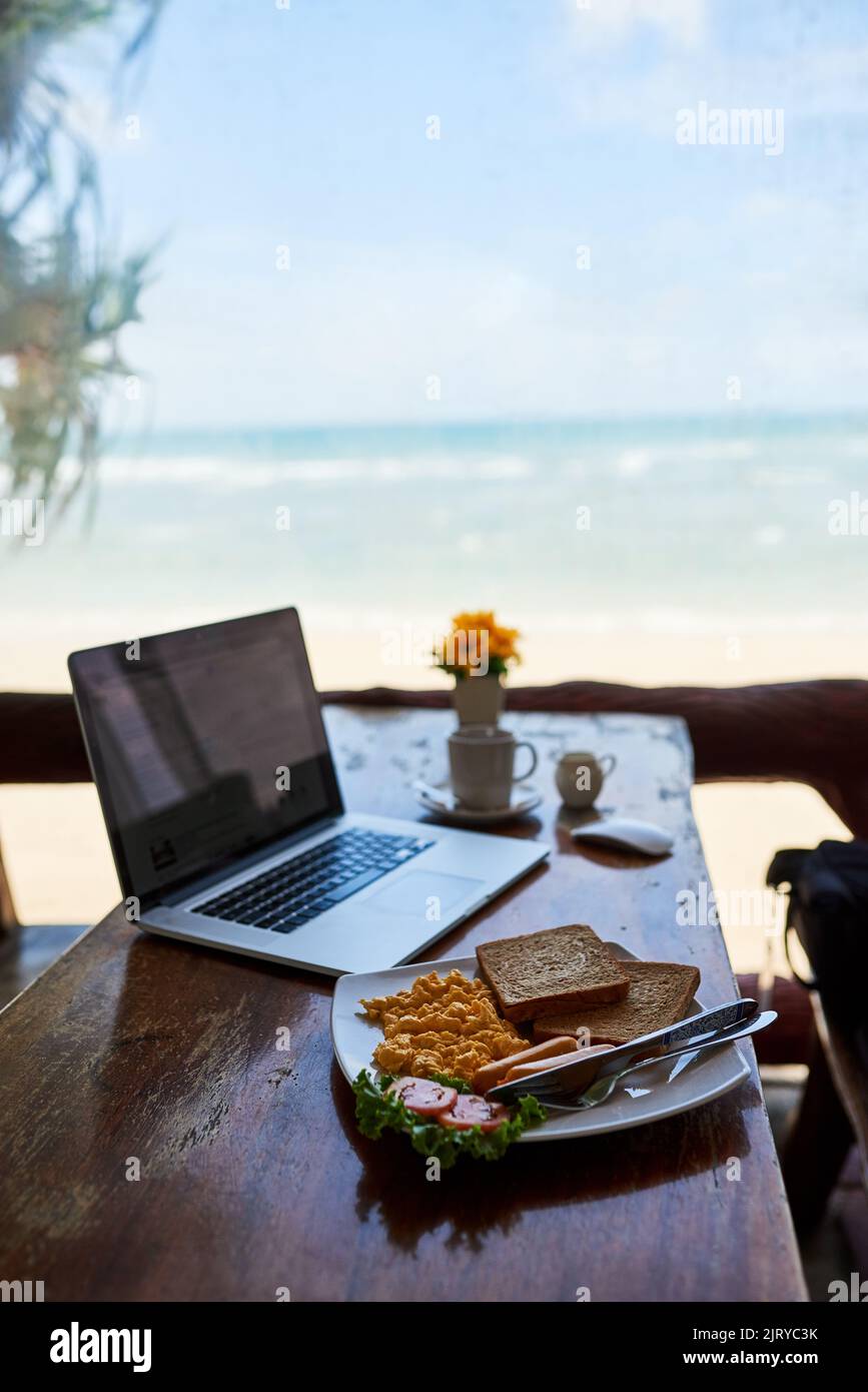 Petit déjeuner à la plage. Ordinateur portable et petit déjeuner fraîchement préparé sur une table avec vue sur la plage en arrière-plan. Banque D'Images
