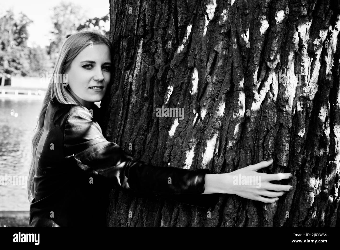 Jolie femme aux longs cheveux blonds, portant une veste en cuir, embrassant un tronc de chêne géant. Concept écologique. Monochrome Banque D'Images