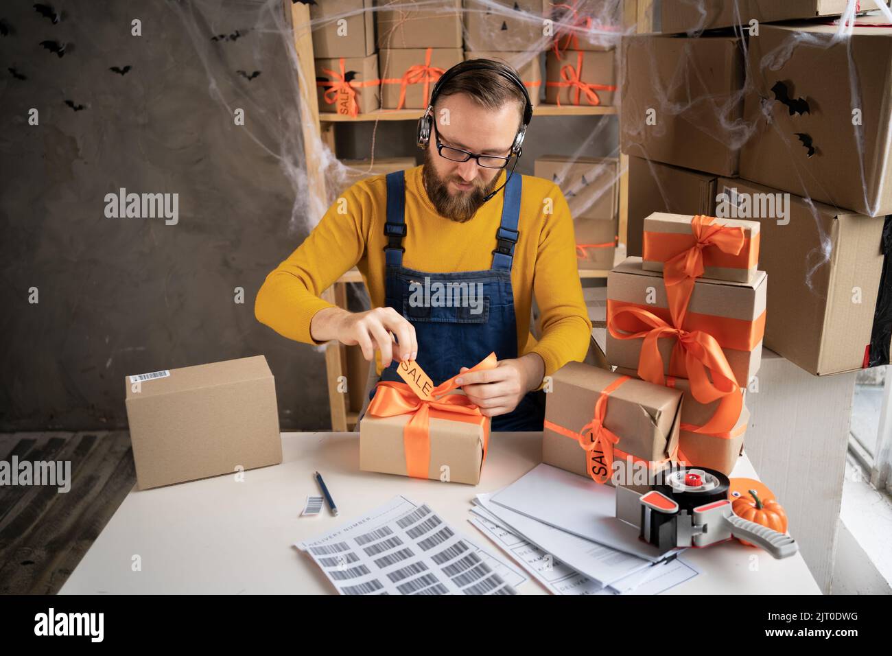 jeune homme barbu en lunettes propriétaire d'affaires assis dans halloween décoré bureau à la maison. Attachez un noeud orange sur la boîte. Vendre des marchandises pour les vacances de tous Banque D'Images