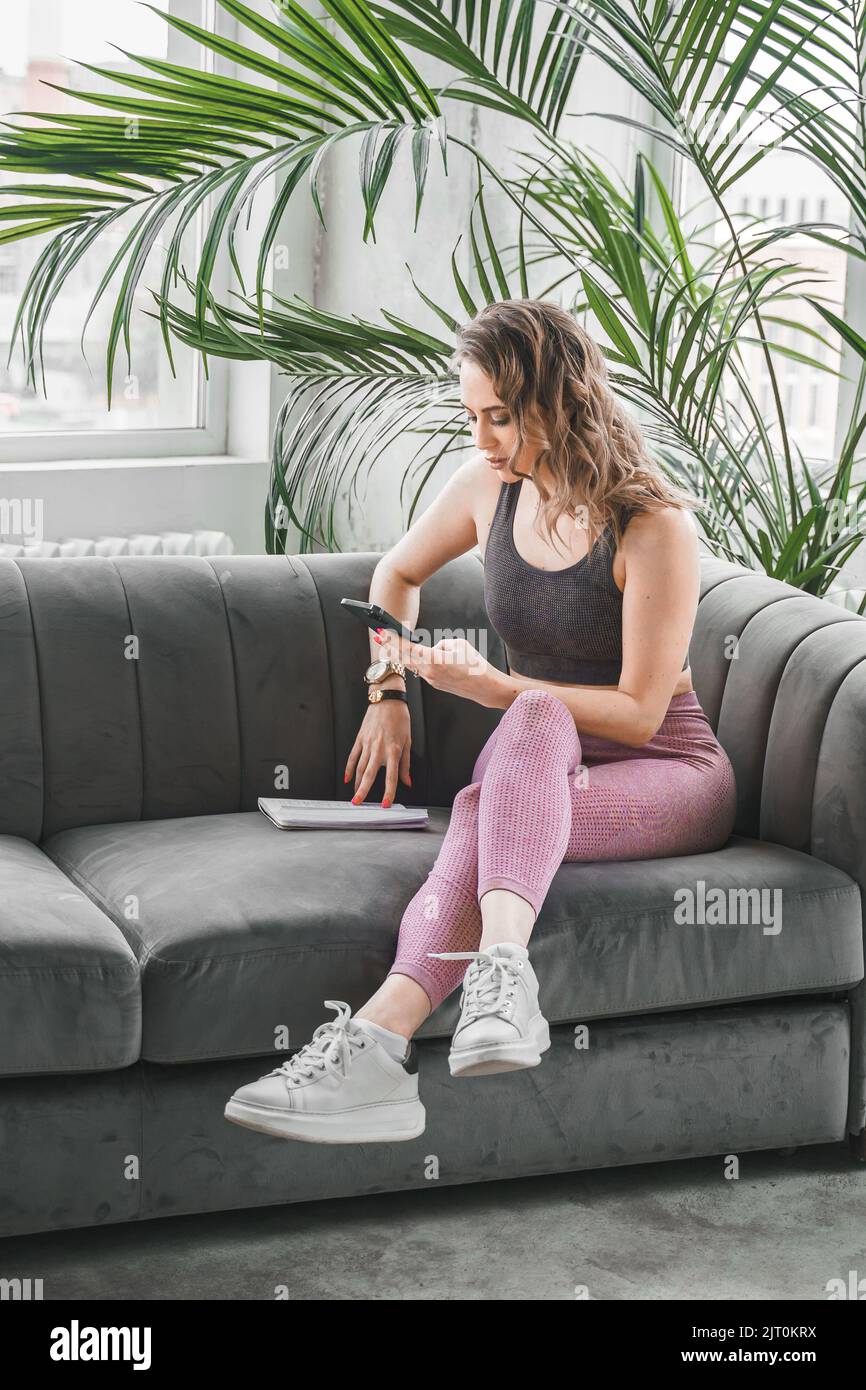 Femme sportive mince entraîneur de yoga posé sur un canapé dans des vêtements de sport avec téléphone mobile. Prendre des notes, planifier. Instructeur de fitness en vêtements de sport Banque D'Images