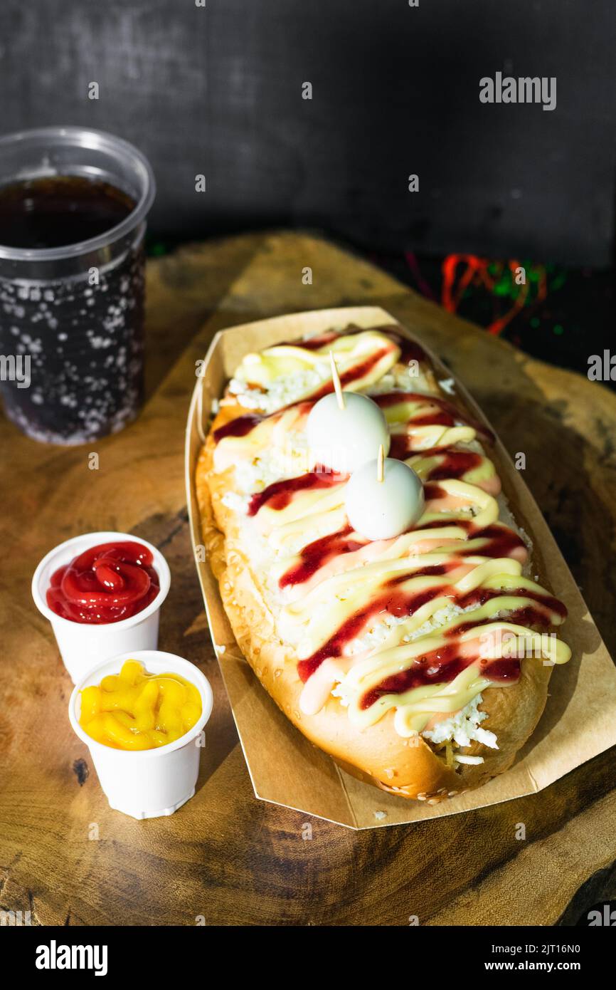 Restauration rapide, hot dog avec sauces, fromage et deux œufs de caille servis sur un plateau en bois et à côté il y a deux tasses en plastique avec ketchup Banque D'Images