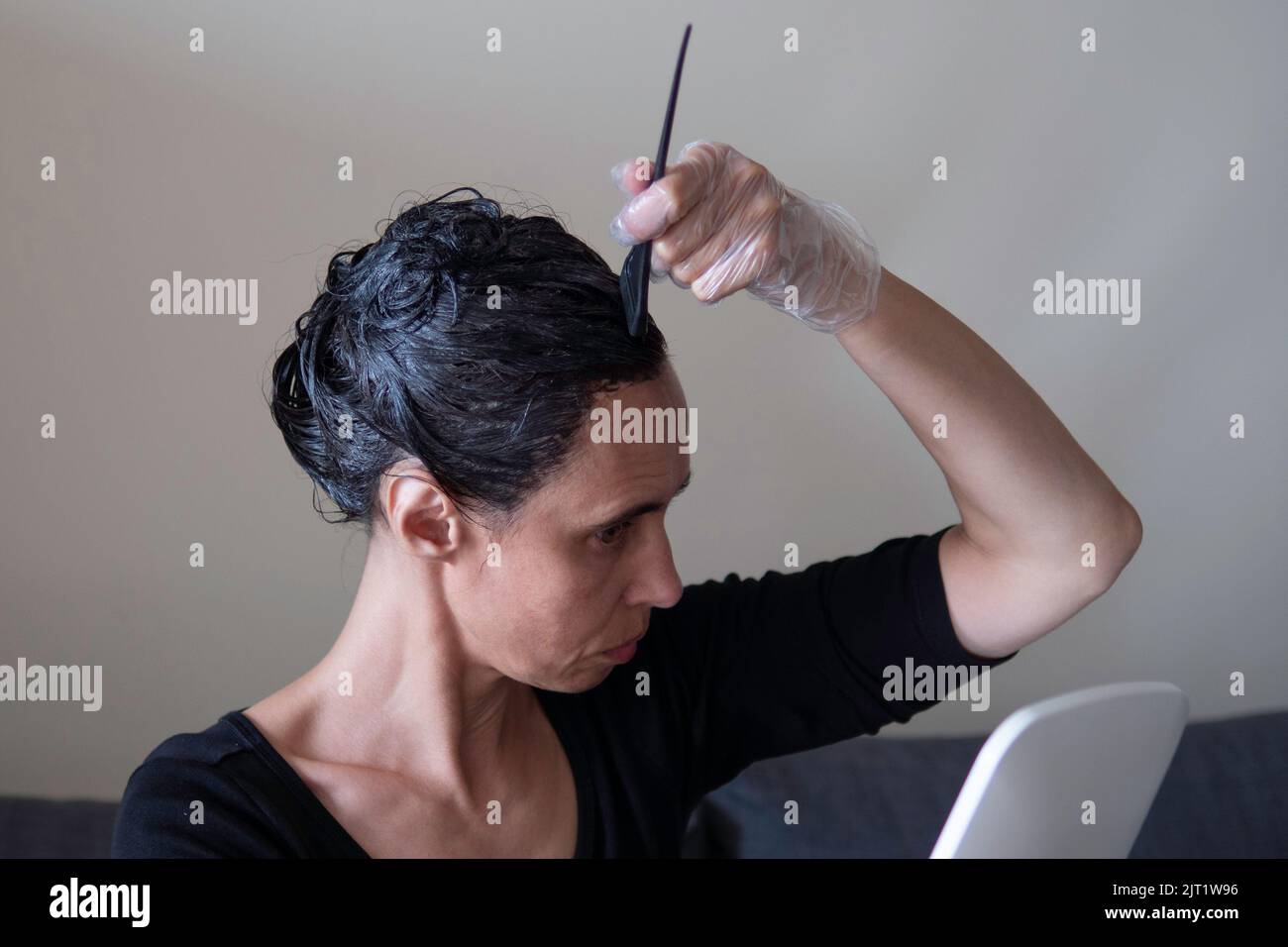 Femme d'âge moyen colorant les cheveux foncés avec des racines grises à la maison. Femme assise sur un canapé et teinture des cheveux à l'aide d'une brosse noire et regardant le mirule Banque D'Images