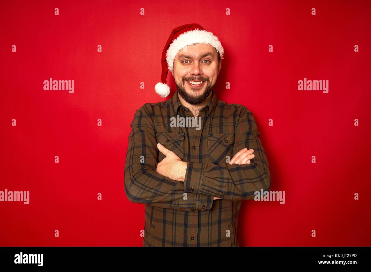 homme souriant en chapeau de nouvel an, vêtements décontractés avec les bras repliés devant isolé arrière-plan de studio rouge Banque D'Images