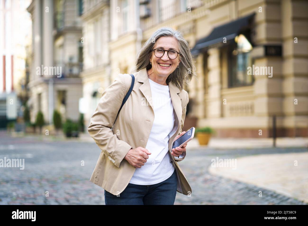 Femme d'affaires élégante et mature. Femme âgée en tenue décontractée tenue habillée tient le téléphone à la main marchant dans la rue du travail ou de retour au bureau. Une femme mature passe du temps libre à voyager dans la ville européenne. Banque D'Images