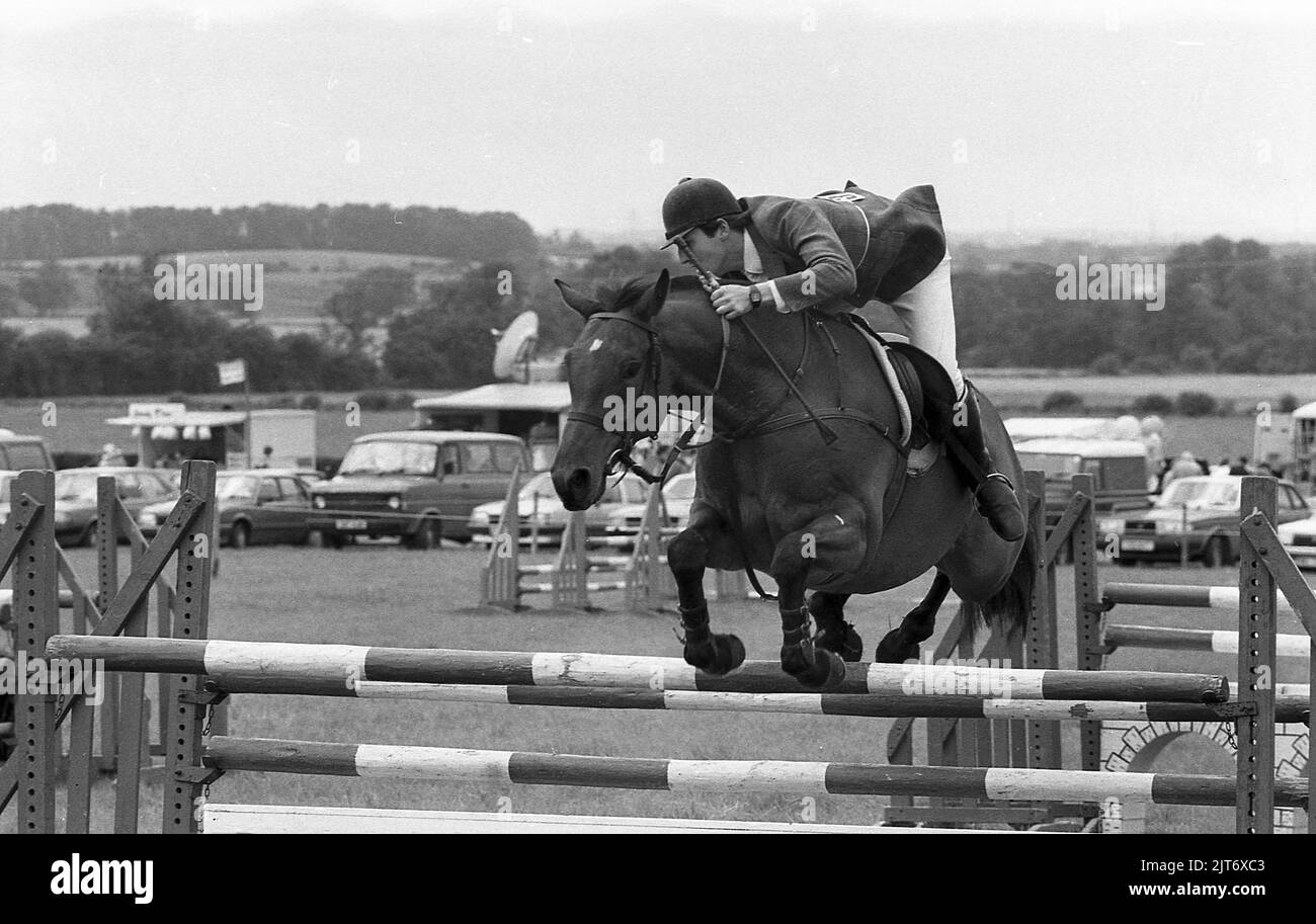 1980s, un showjump masculin sur un cheval sautant une clôture à un spectacle de comté, Yorkshire, Angleterre, Royaume-Uni. Le saut de spectacle est un événement équestre où le cavalier et le cheval tentent de sauter sur toutes les clôtures sur un parcours, sans les frapper et dans un délai défini. Banque D'Images