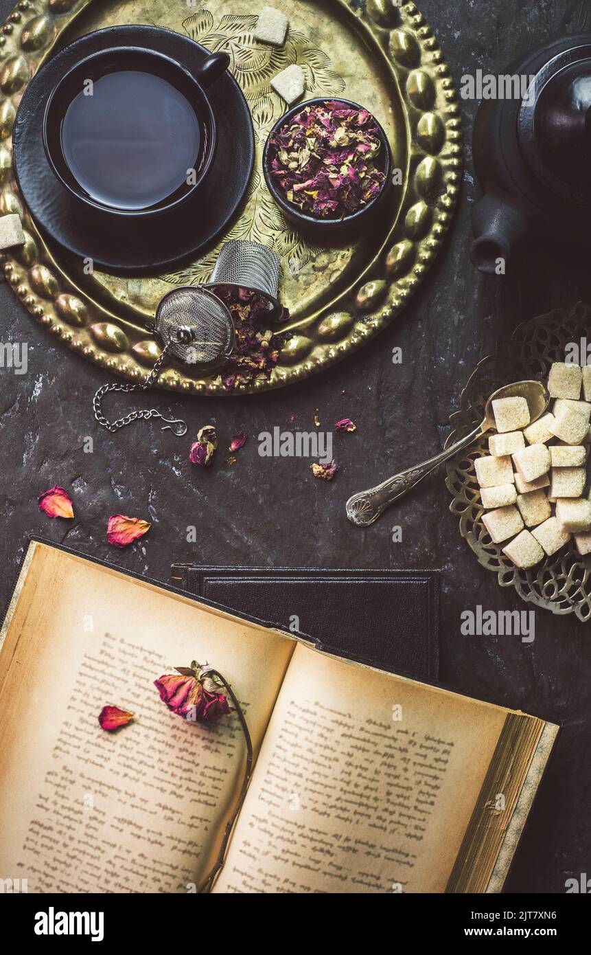 Encore la vie avec une tasse de thé lieu avec des fleurs sèches, sucre, thé noir pot et livre vintage sur fond rustique sombre Banque D'Images