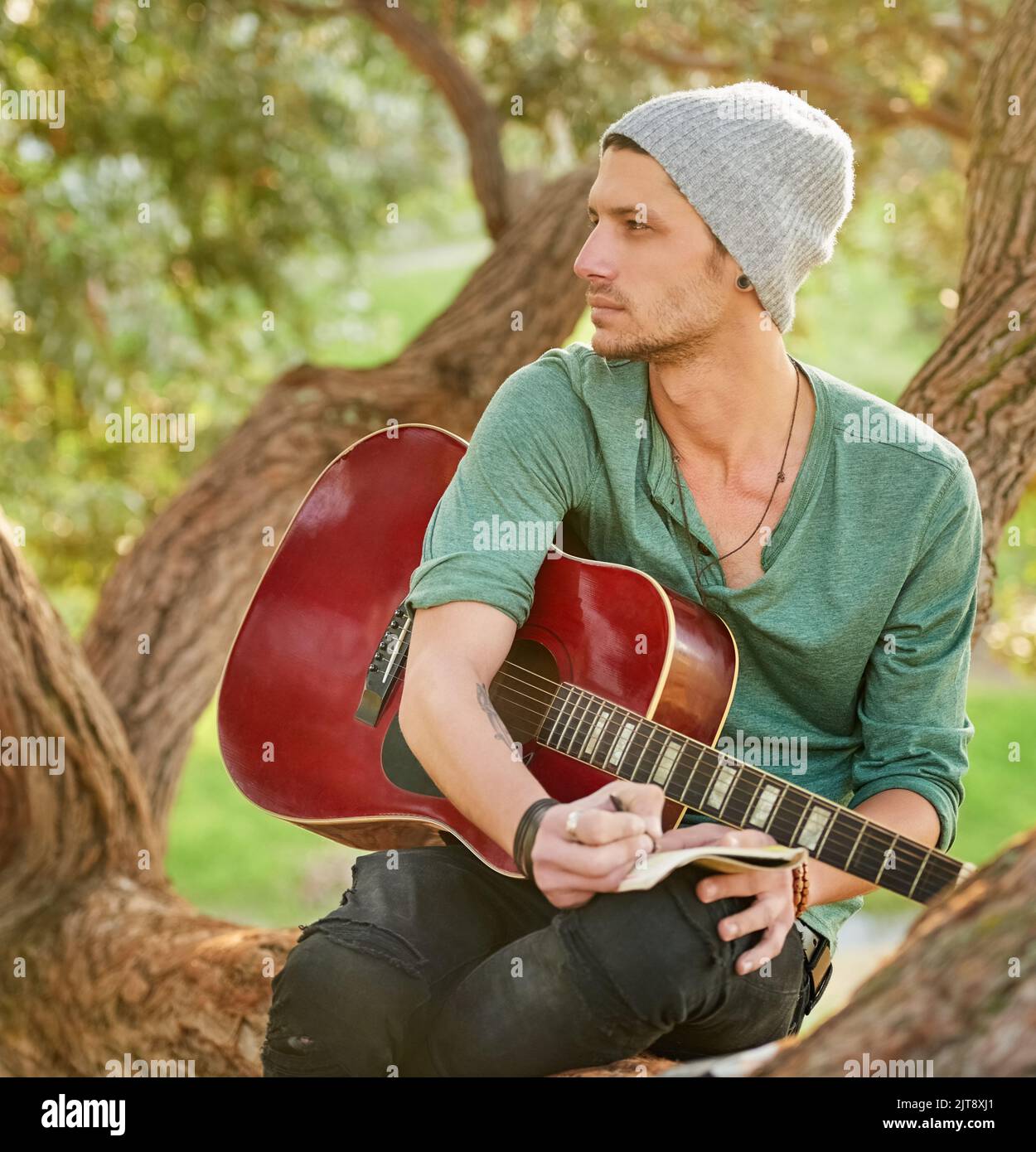 S'inspirer musicalement de la nature. Un beau jeune homme assis dans un parc avec une guitare écrivant une chanson sur un bloc-notes. Banque D'Images