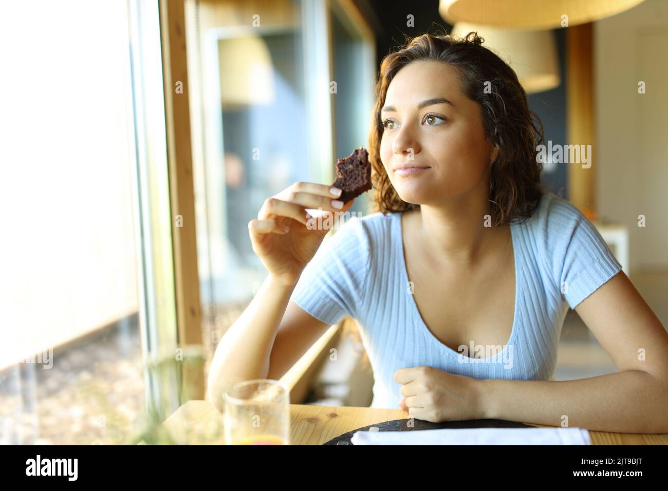 Femme tenant et mangeant un cupcake au chocolat assis dans un restaurant regardant par une fenêtre Banque D'Images