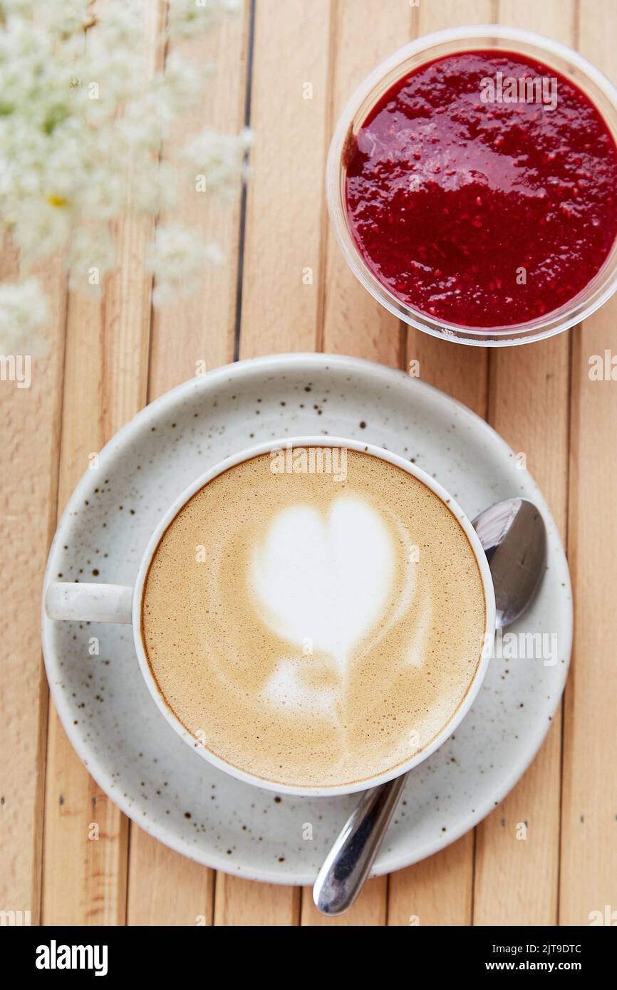 Tasse de cappuccino blanc en céramique au coeur, dessert de confiture de framboises - apéritif, petit déjeuner léger sur la terrasse du café avec avant-plan flou de fleurs de camomille. Gros plan de la nourriture. Photo verticale. Banque D'Images