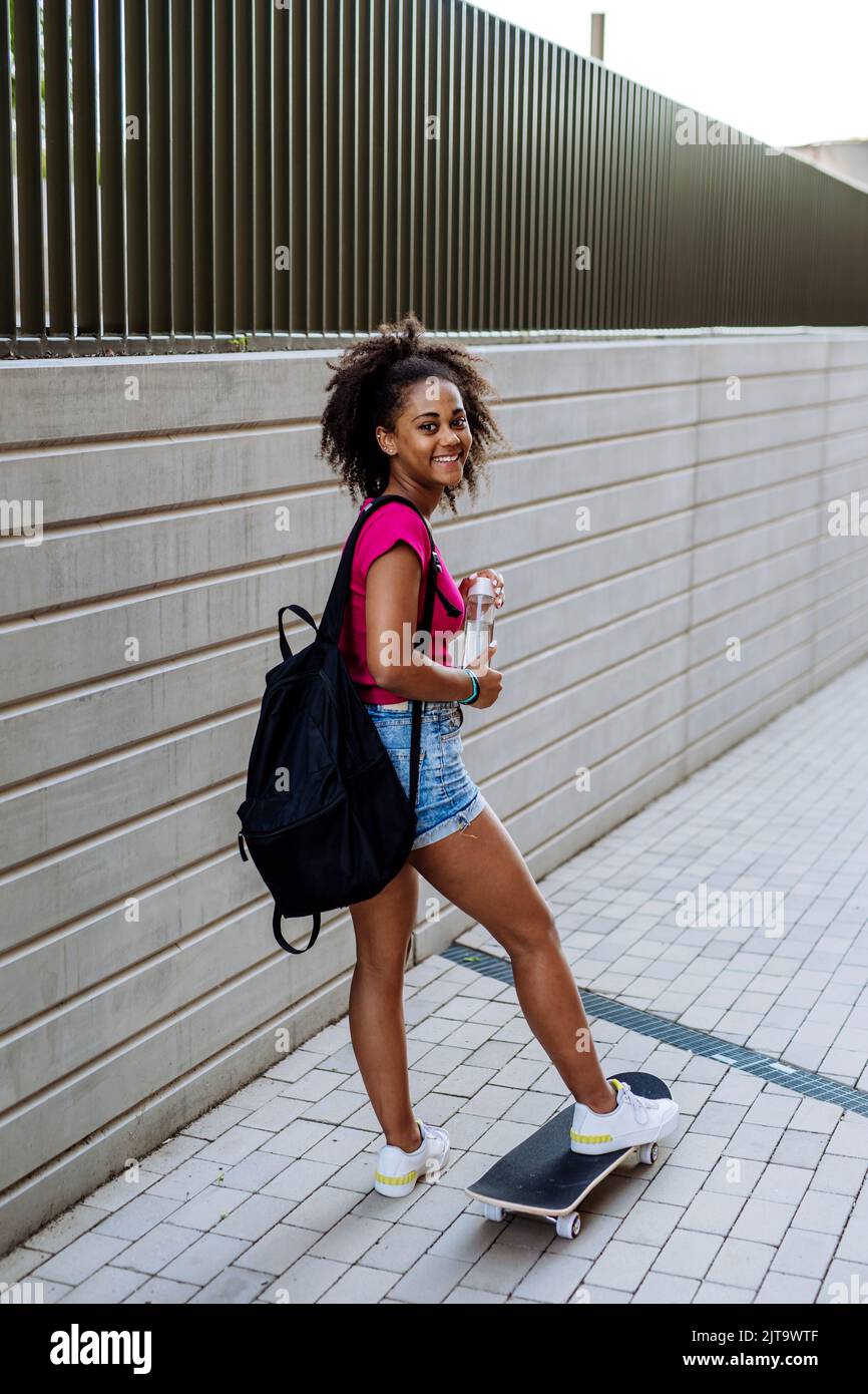 Adolescente multiraciale avec sac à dos à la planche à roulettes, pendant la journée d'été. Banque D'Images