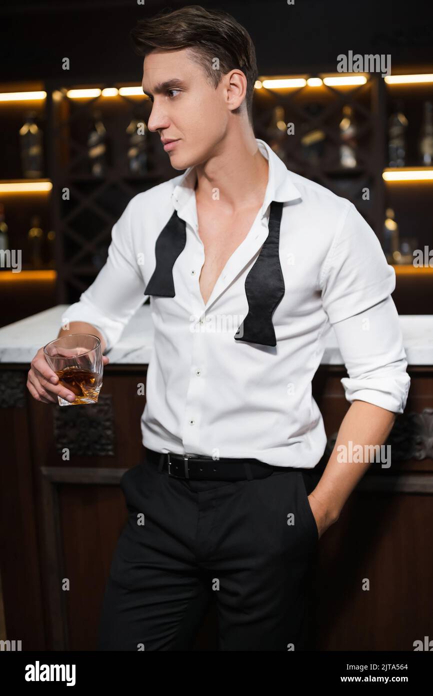 Homme élégant en chemise tenant un verre de whisky près du bar le soir Banque D'Images