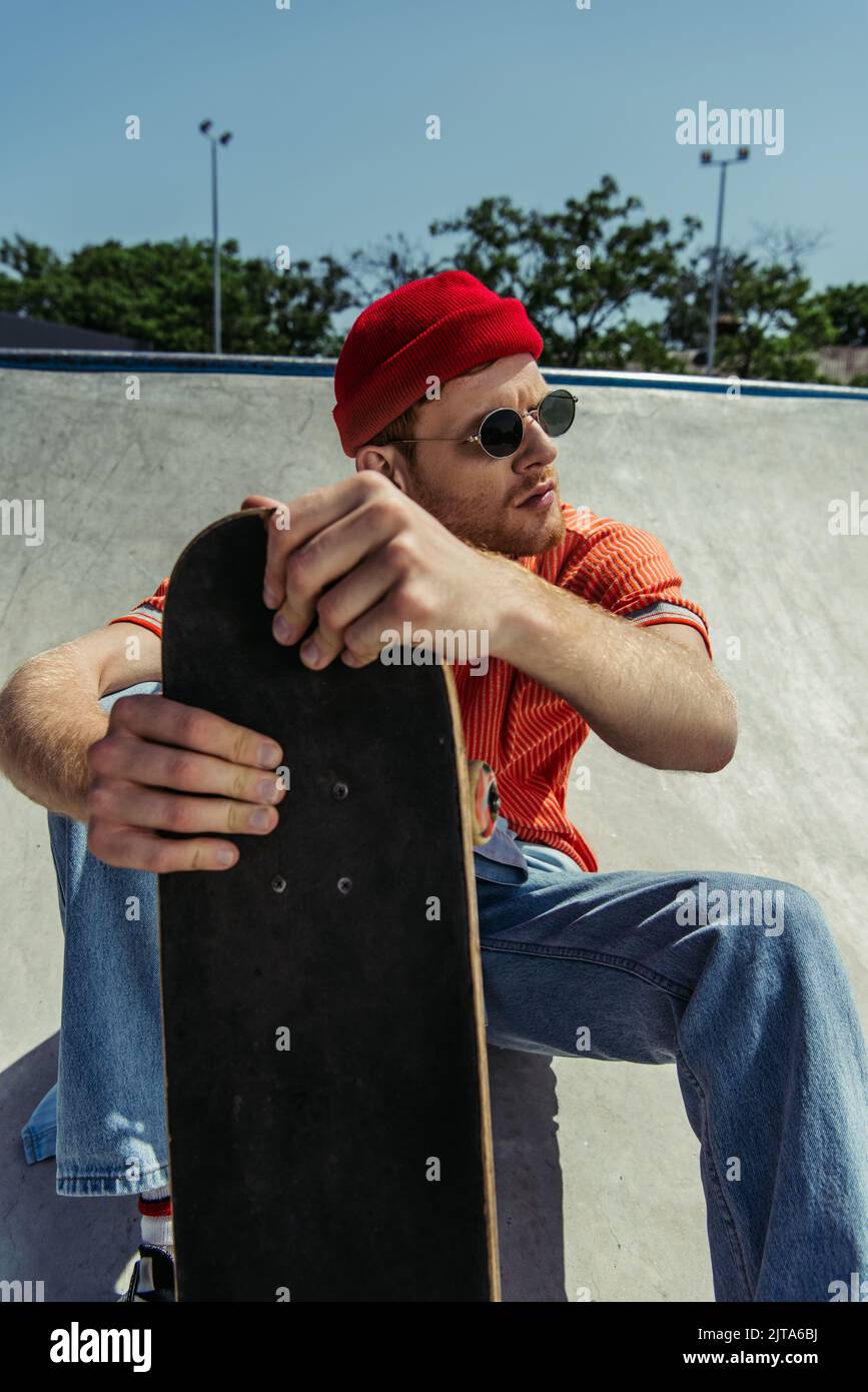 jeune homme tendance dans des lunettes de soleil assis avec un skateboard et regardant loin Banque D'Images