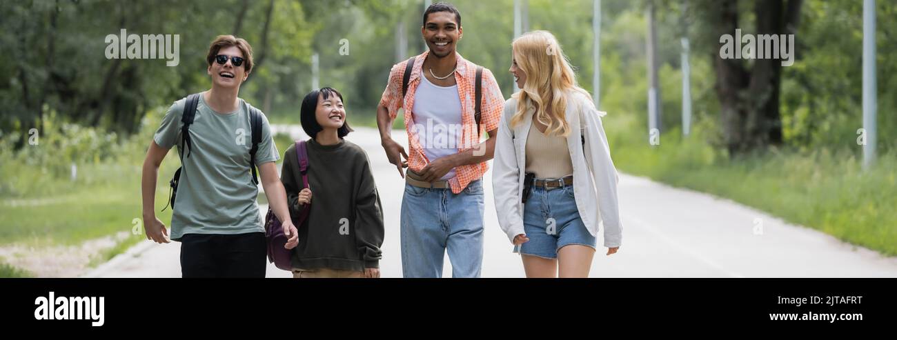 jeunes et joyeux amis interraciaux marchant sur la route près de la forêt, bannière Banque D'Images