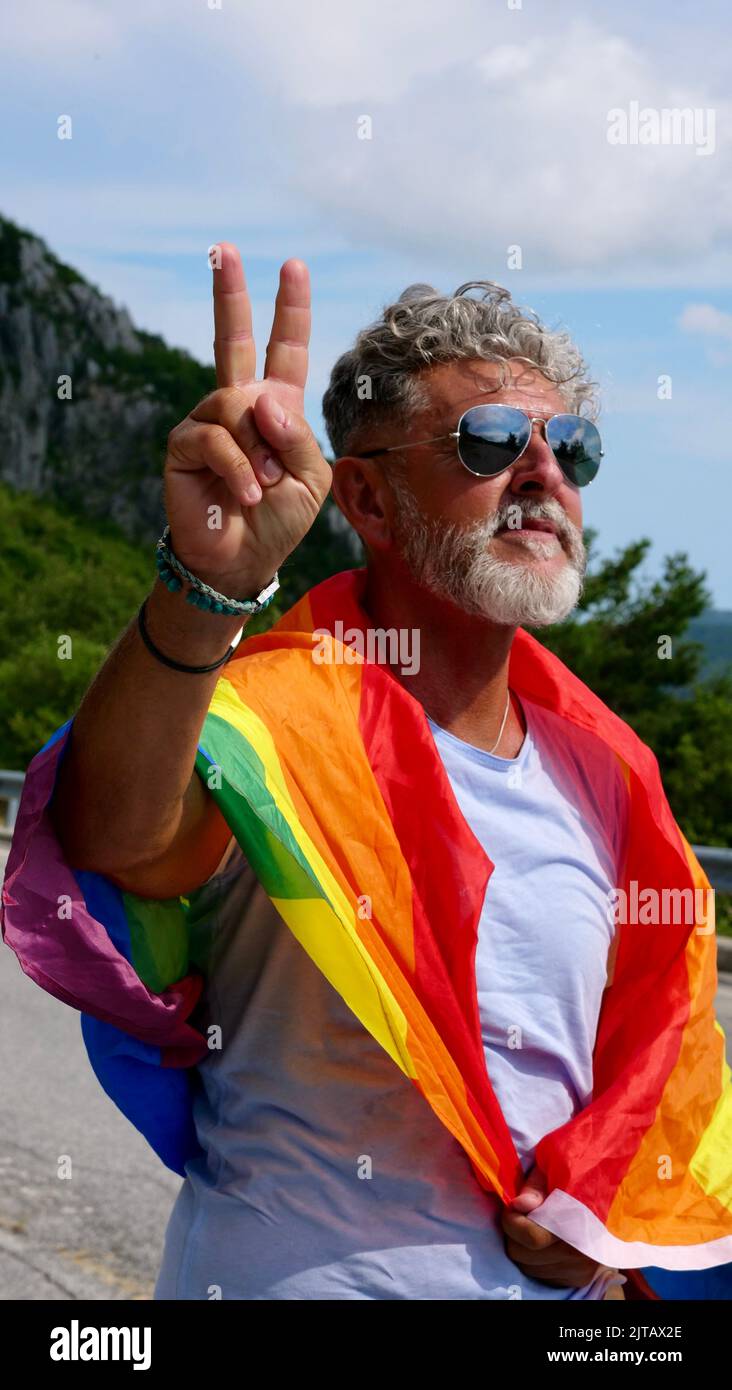 Portrait d'un homme caucasien âgé âgé âgé à cheveux gris bisexualité avec une barbe et des lunettes de soleil avec un drapeau de paix LGBTQIA arc-en-ciel dans les montagnes. Célèbre le mois de la fierté, le Rainbow Flag Day, la parade des gays Banque D'Images