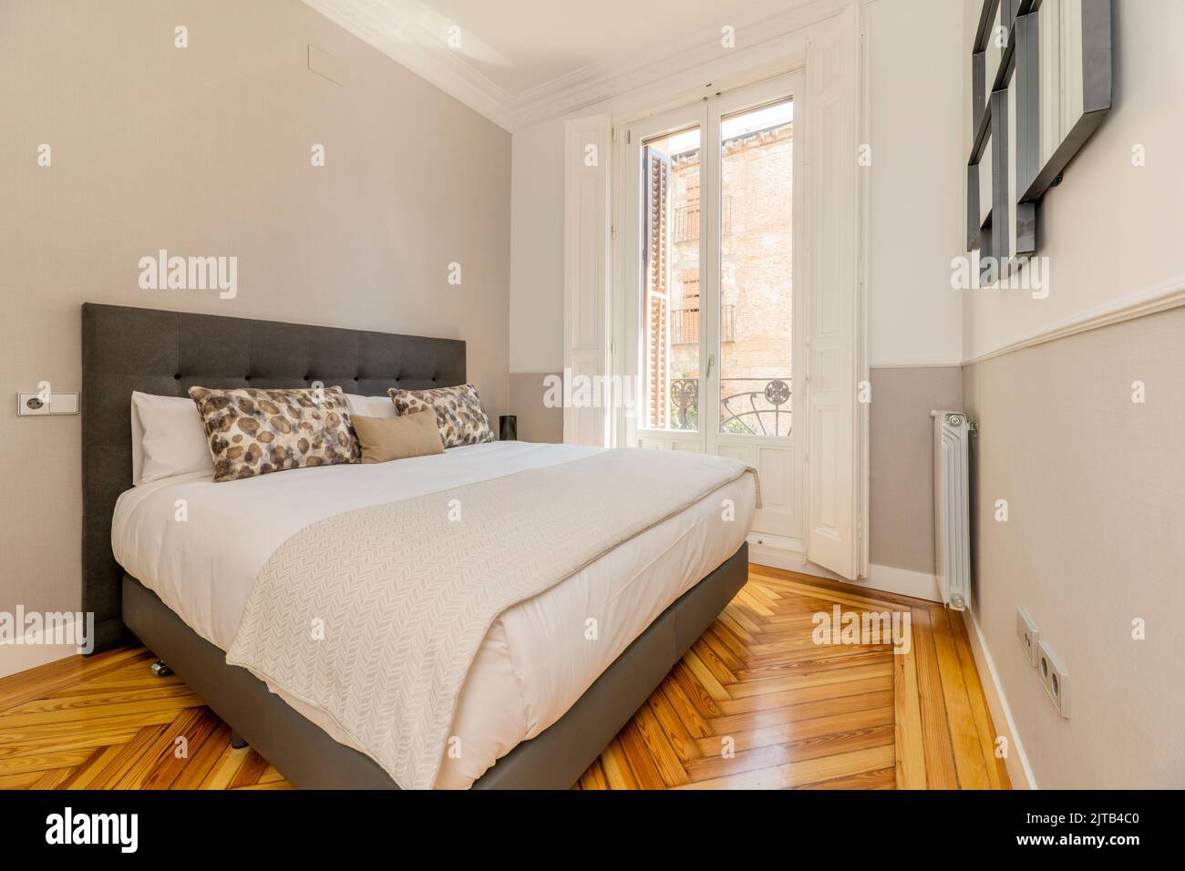 Chambre avec tête de lit rembourrée en tissu gris, coussins et oreillers assortis, grand balcon avec fenêtres et volets blancs, couvertures assorties, origi Banque D'Images