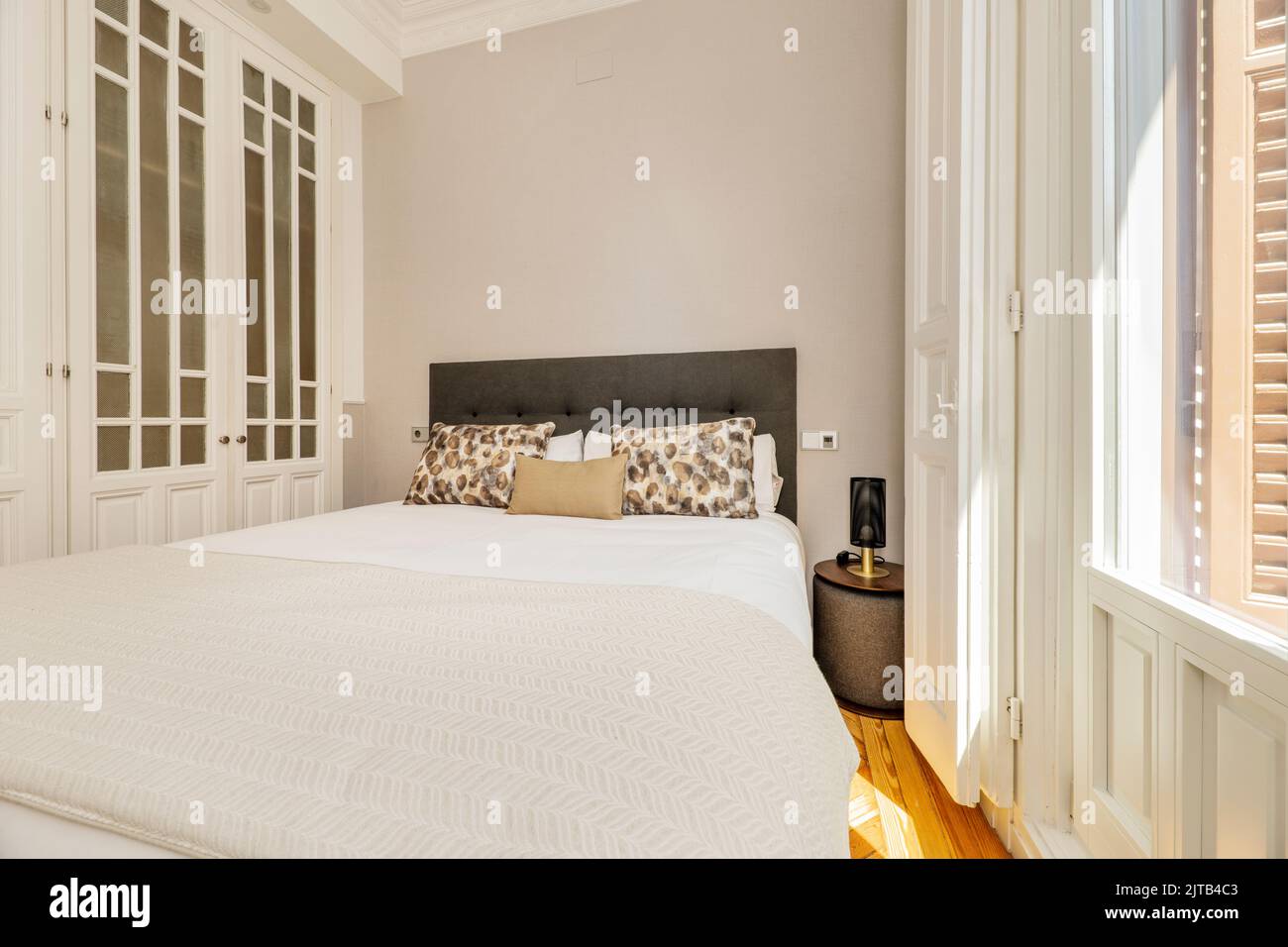 Chambre avec tête de lit rembourrée en tissu gris, coussins et oreillers assortis, grand balcon avec fenêtres et volets blancs et armoire intégrée Banque D'Images