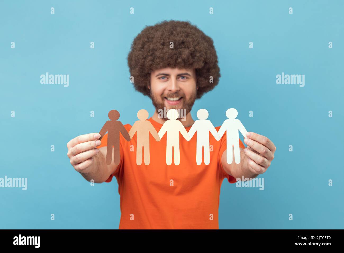 Portrait d'un homme heureux avec une coiffure afro portant un T-shirt orange tenant des gens en papier tenant les mains, différentes cultures, le travail d'équipe et le partenariat. Studio d'intérieur isolé sur fond bleu. Banque D'Images