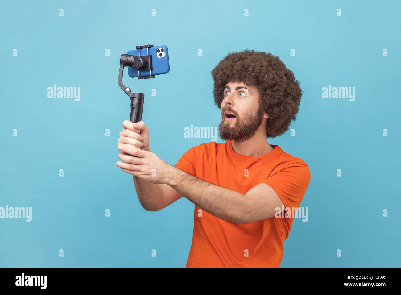 Portrait de l'homme surpris avec la coiffure afro portant le T-shirt orange debout avec les grands yeux et tenant Steadicam pour prendre le selfie. Studio d'intérieur isolé sur fond bleu. Banque D'Images Portrait de l'homme surpris avec la coiffure afro portant le T-shirt orange debout avec les grands yeux et tenant Steadicam pour prendre le selfie. Studio d'intérieur isolé sur fond bleu. Banque D'Images