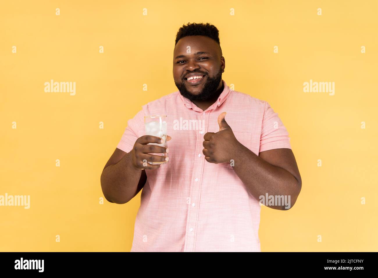 Portrait d'un homme heureux souriant et souriant, portant une chemise rose, a soif, tenant un verre d'eau froide fraîche avec de la glace, montrant le pouce vers le haut. Studio d'intérieur isolé sur fond jaune. Banque D'Images