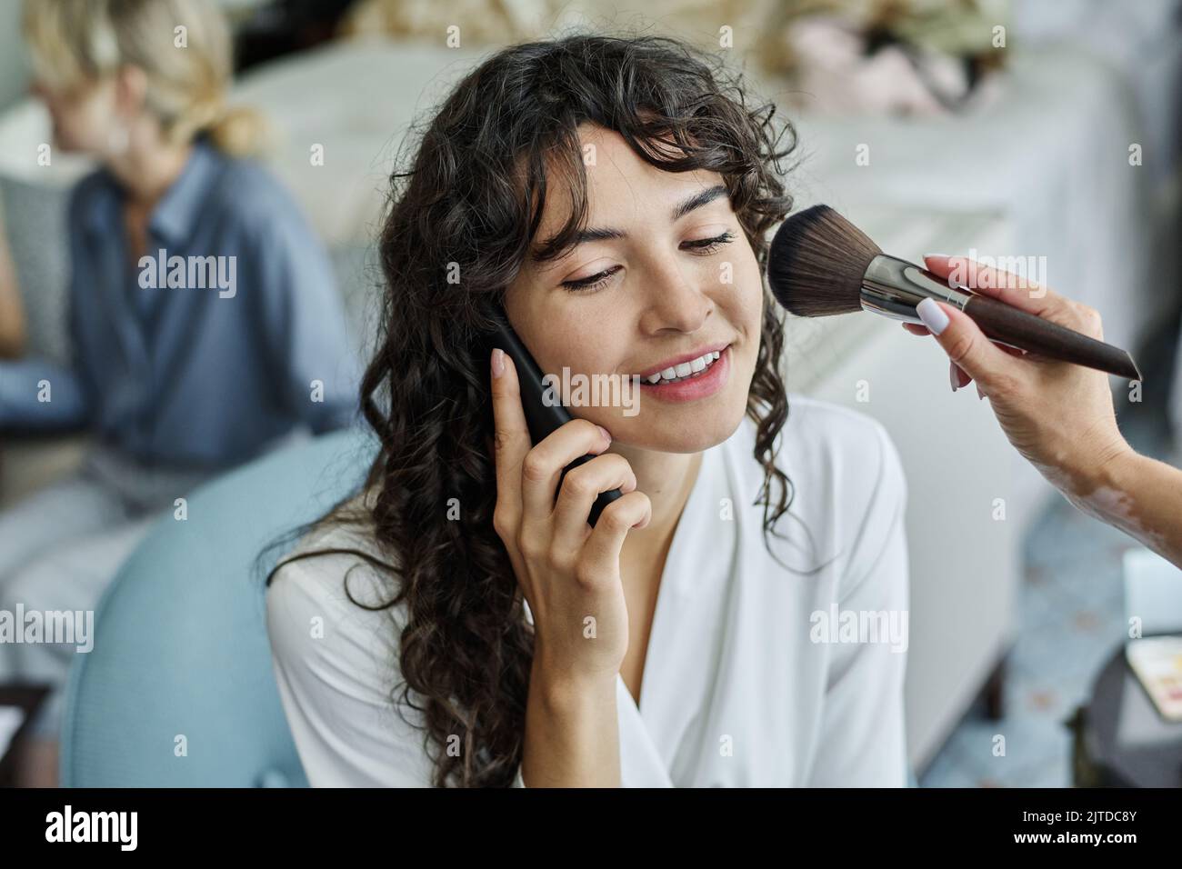 Bonne jeune mariée avec de longs cheveux ondulés foncés parlant sur le smartphone tandis que maquilleuse avec brosse appliquant de la poudre sur sa joue Banque D'Images
