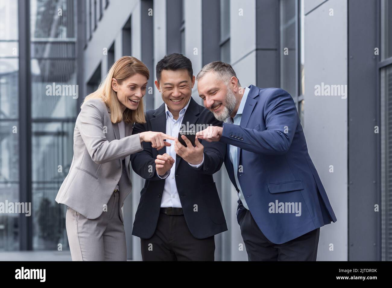 Groupe d'affaires diversifié, trois hommes et femmes travaillant à l'extérieur du bâtiment de bureau souriant gaiement et heureux de regarder la vidéo au téléphone Banque D'Images