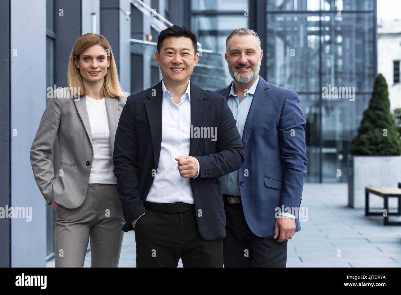 Équipe d'affaires diversifiée réussie, trois employés souriant et regardant la caméra, équipe de rêve avec un patron asiatique à l'extérieur du bâtiment de bureau, des collègues dans des costumes d'affaires Banque D'Images