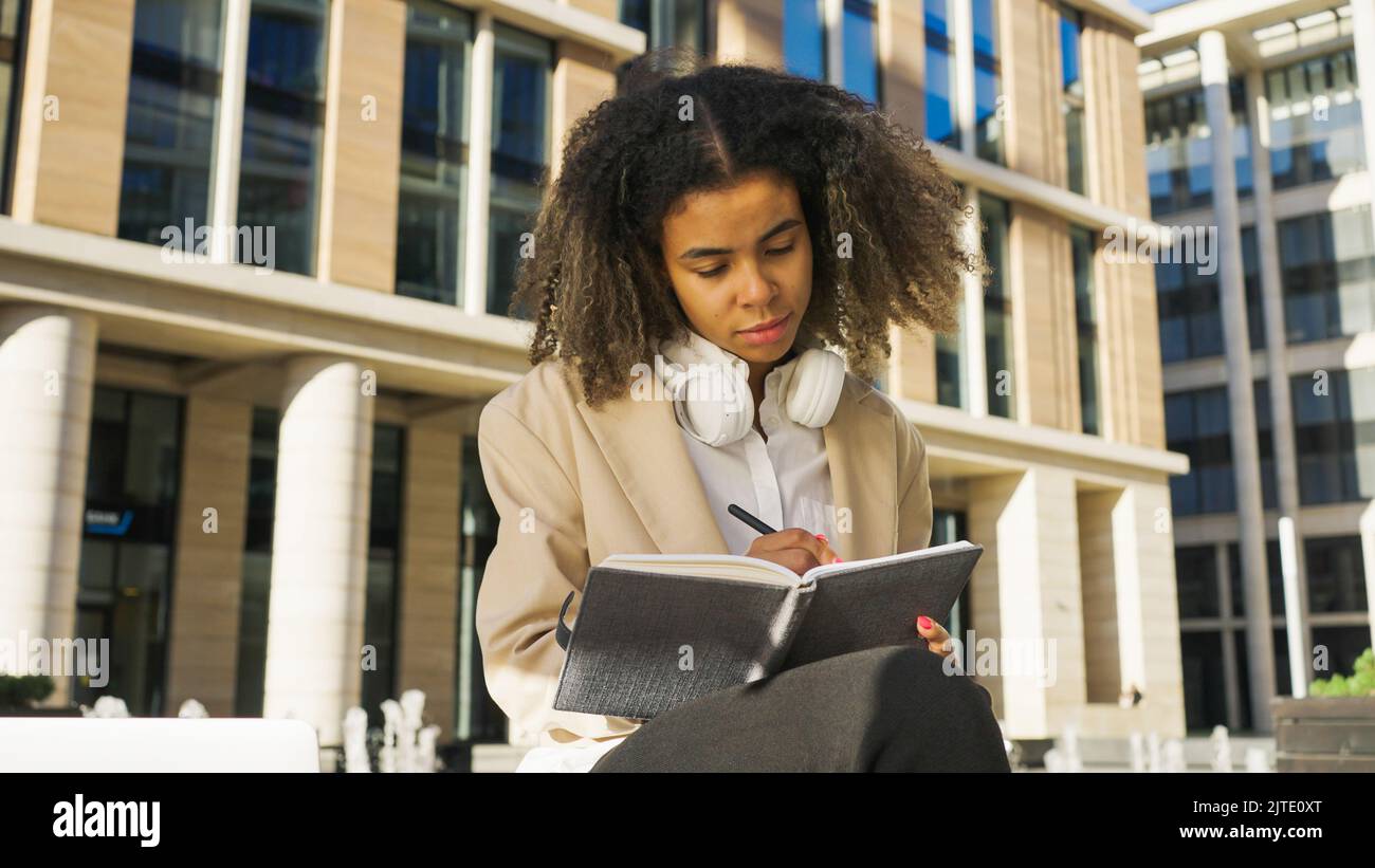 Une femme afro-américaine écrit des plans dans un carnet papier avec une expression réfléchie. Une jeune femme d'affaires pense à la future start-up en train de prendre des notes par immeuble de bureaux Banque D'Images