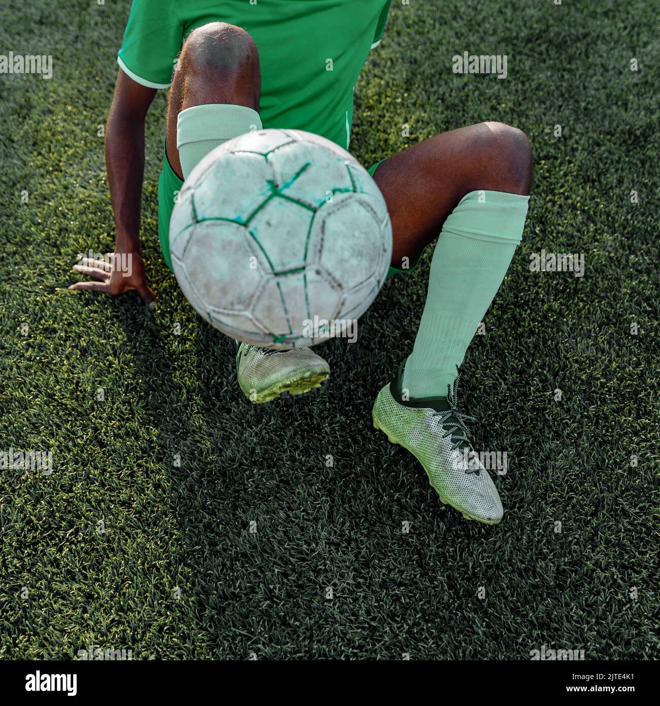 Homme de couleur à la peau sombre non reconnaissable, dans un uniforme de football vert tenant un ballon de football tout en étant allongé sur le terrain de football Banque D'Images