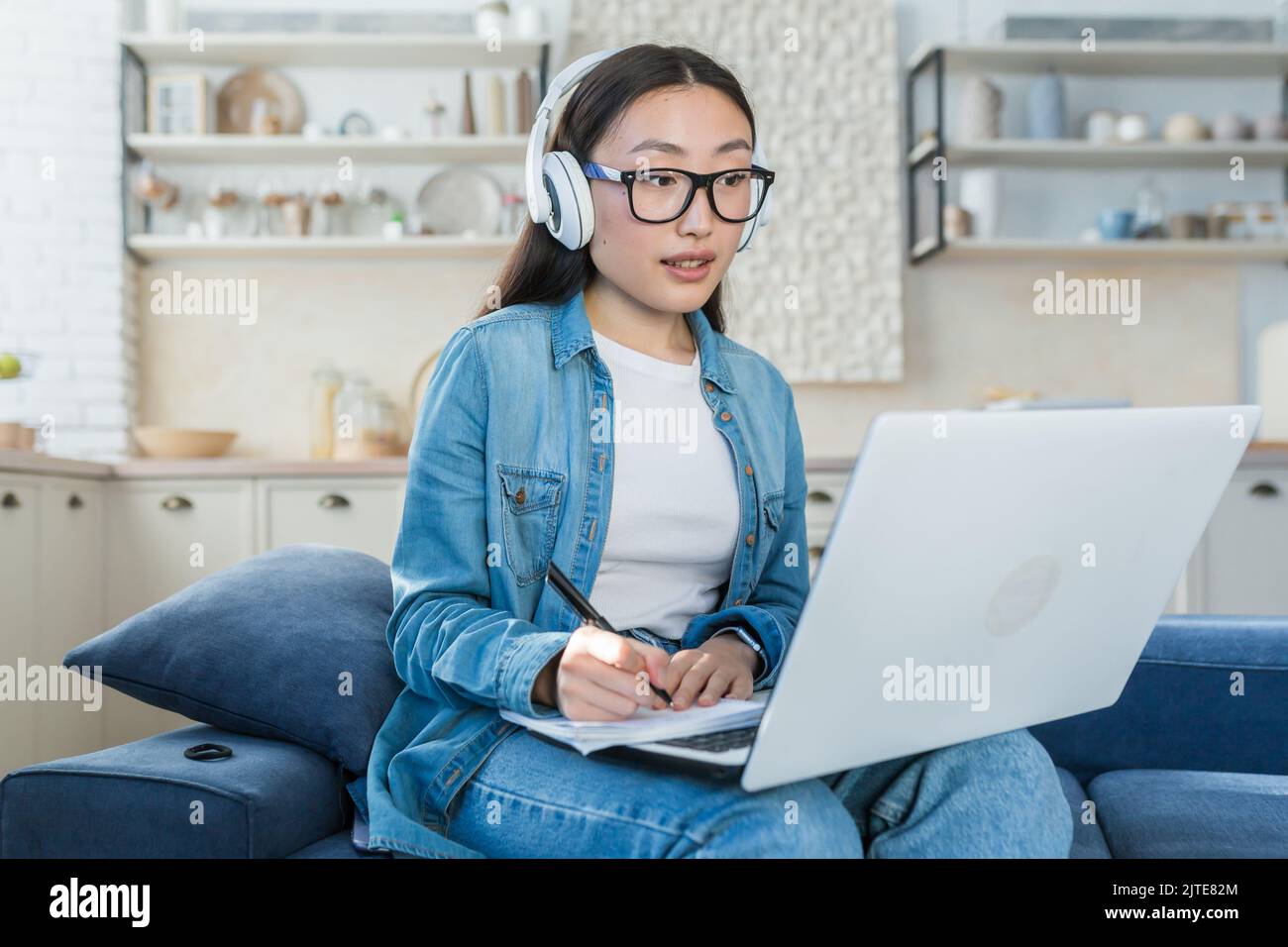 Jeune femme asiatique qui étudie à distance à la maison, étudiante qui écoute des cours en ligne, prend des notes, s'asseyant sur un canapé dans le salon à l'aide d'un casque et d'un ordinateur portable Banque D'Images