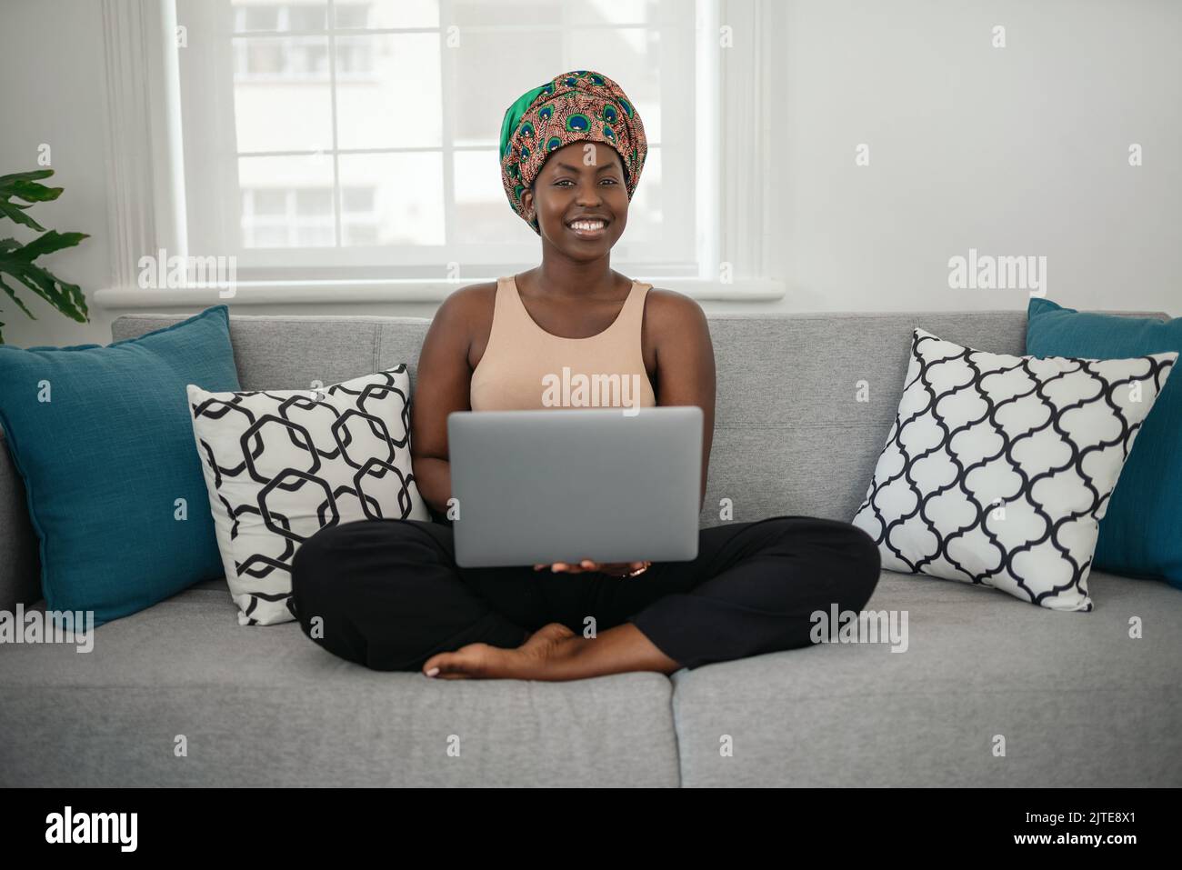 Une jeune femme afro-américaine souriante assise avec ses jambes croisées sur son canapé à la maison à l'aide d'un ordinateur portable et regardant dans l'appareil photo Banque D'Images