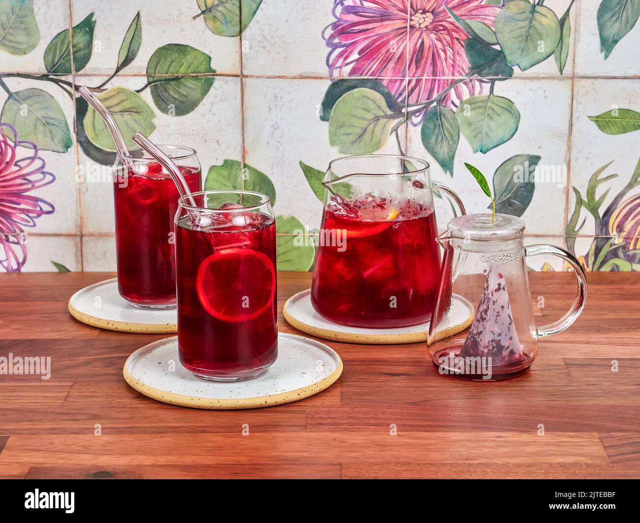Thé glacé à la framboise dans une théière en verre spéciale, avec deux verres à thé glacé et des pailles borosilicates, sur une table en noyer et des carreaux de céramique en arrière-plan. Banque D'Images