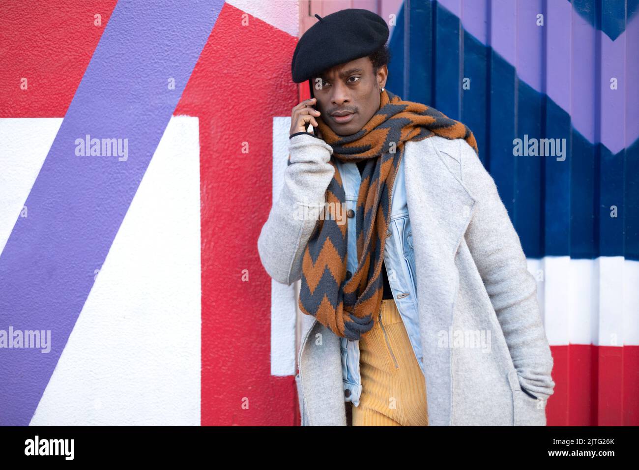 Jeune homme afro-américain adulte parlant au téléphone avec un geste sérieux. Il se penche contre un mur coloré. Banque D'Images