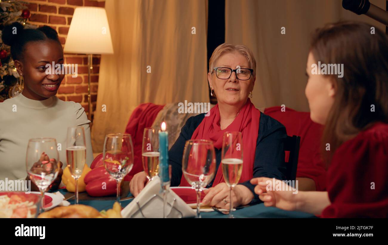Bonne femme senior assise à la table du dîner de Noël avec des membres de la famille tout en regardant l'appareil photo. Personne âgée festive célébrant des vacances traditionnelles d'hiver avec des proches à la maison. Banque D'Images
